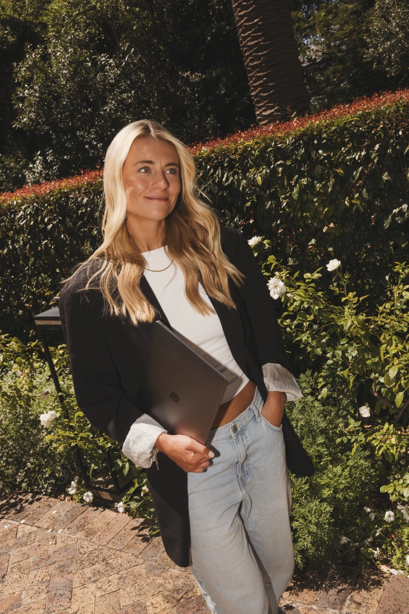 Eva Huese in een tuin met een zwart colbert en haar laptop onder de arm, ze kijkt opzij met een lichte glimlach