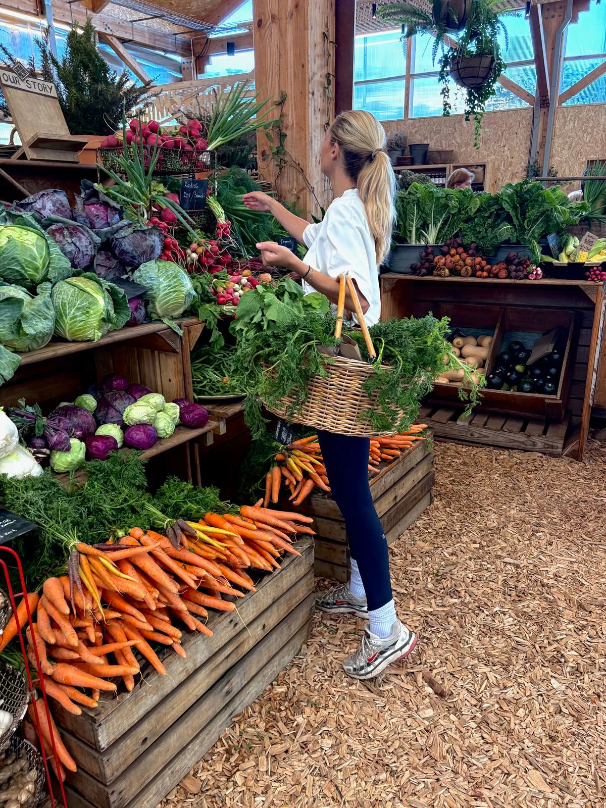 Eva Huese kiest groenten bij een marktkraam, met een rieten mand vol groen aan haar arm